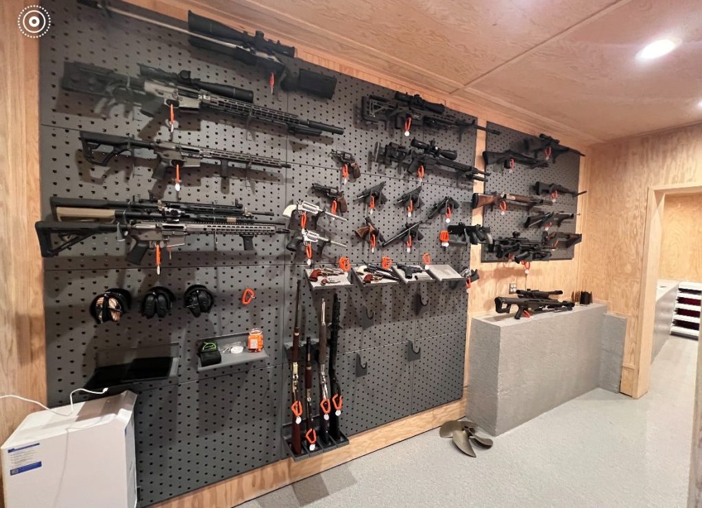 Wall-mounted gun rack displaying rifles, shotguns, and firearms with safety locks in a secured storage room.